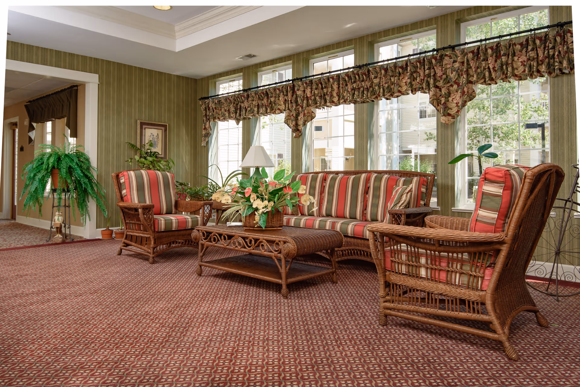 A cozy sitting area with wicker furniture including a sofa and two armchairs with red and green striped cushions. A wicker coffee table with a floral arrangement is in the center. The room has large windows with floral valances, green striped wallpaper, and a patterned carpet. There are several potted plants and a lamp on a side table.