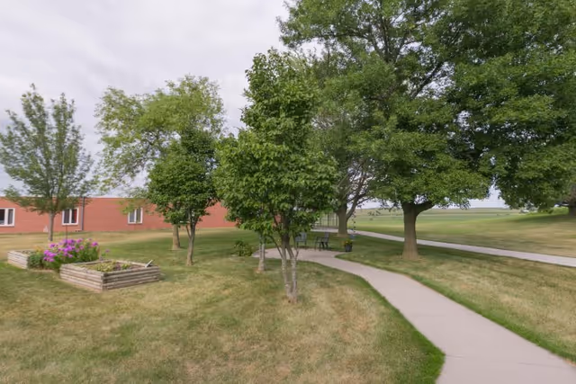Outdoor area of Colonial Manor Of Randolph featuring a curved concrete pathway surrounded by green grass, several leafy trees, a wooden raised garden bed with flowers, and a brick building in the background under a cloudy sky.