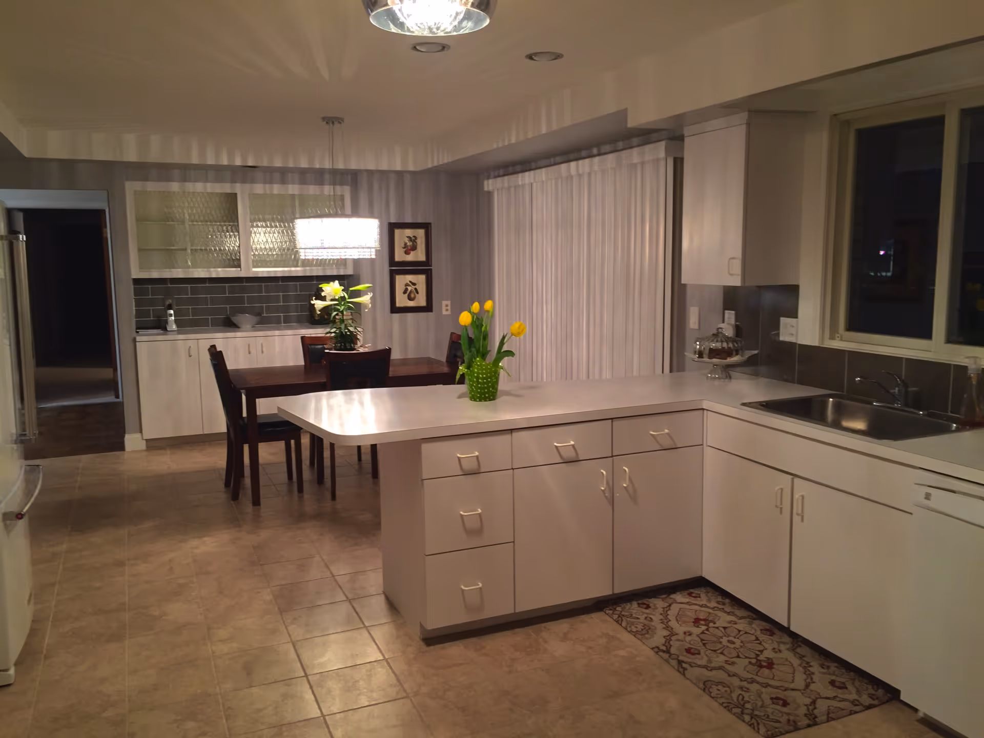 A clean and well-lit kitchen and dining area with white cabinets, a countertop island with a green vase holding yellow tulips, a stainless steel sink, and a dining table with chairs and a hanging light fixture above it. The floor is tiled, and there are framed pictures on the wall near the dining area.
