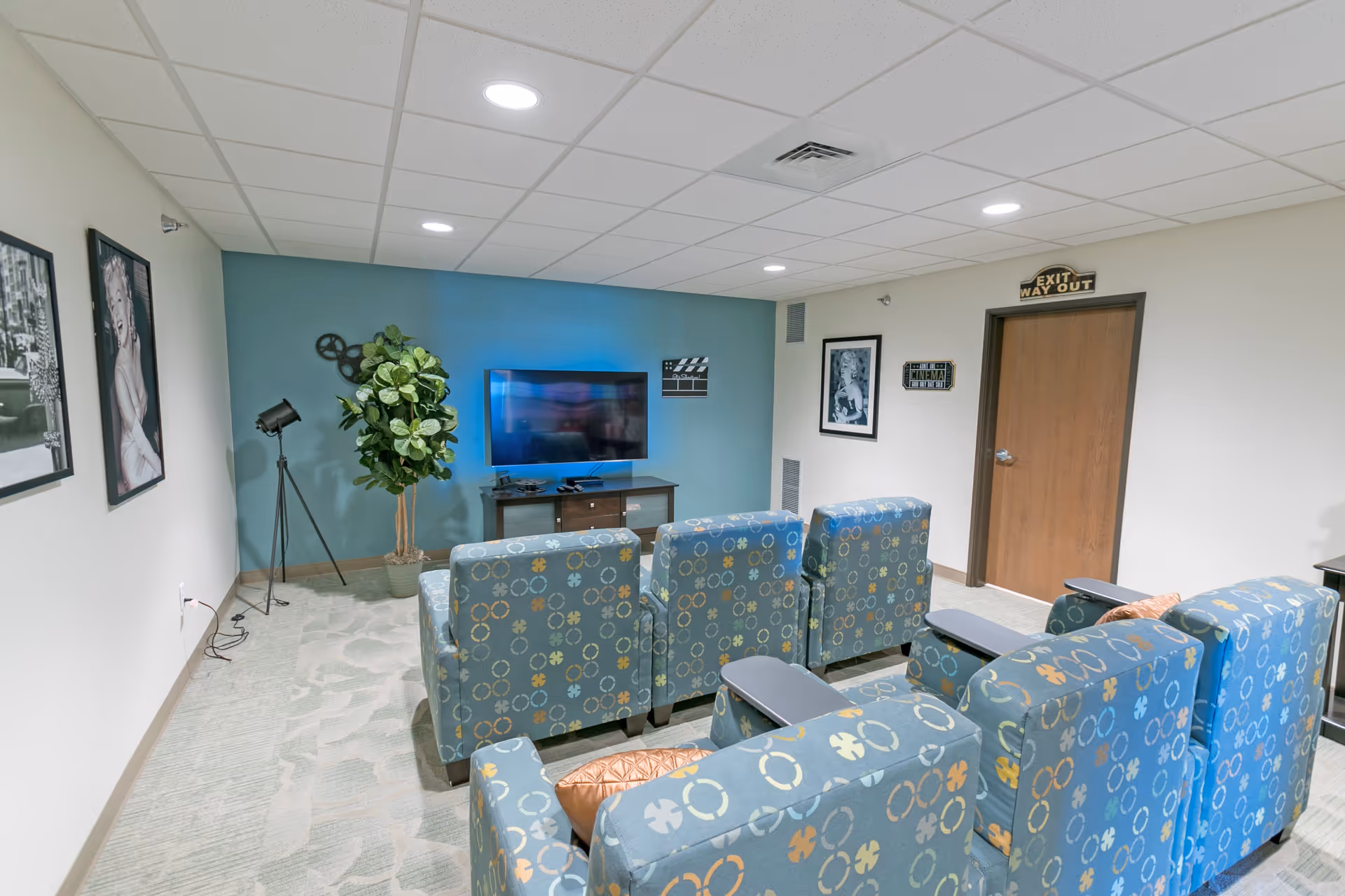 A small movie theater room with six patterned blue armchairs arranged in two rows facing a flat-screen TV mounted on a teal accent wall. The room has a beige carpet with a subtle pattern, white ceiling tiles with recessed lighting, and framed black-and-white photos on the walls. There is a potted plant and a vintage-style spotlight lamp near the TV. A wooden door with an 'EXIT WAY OUT' sign is visible on the right side.