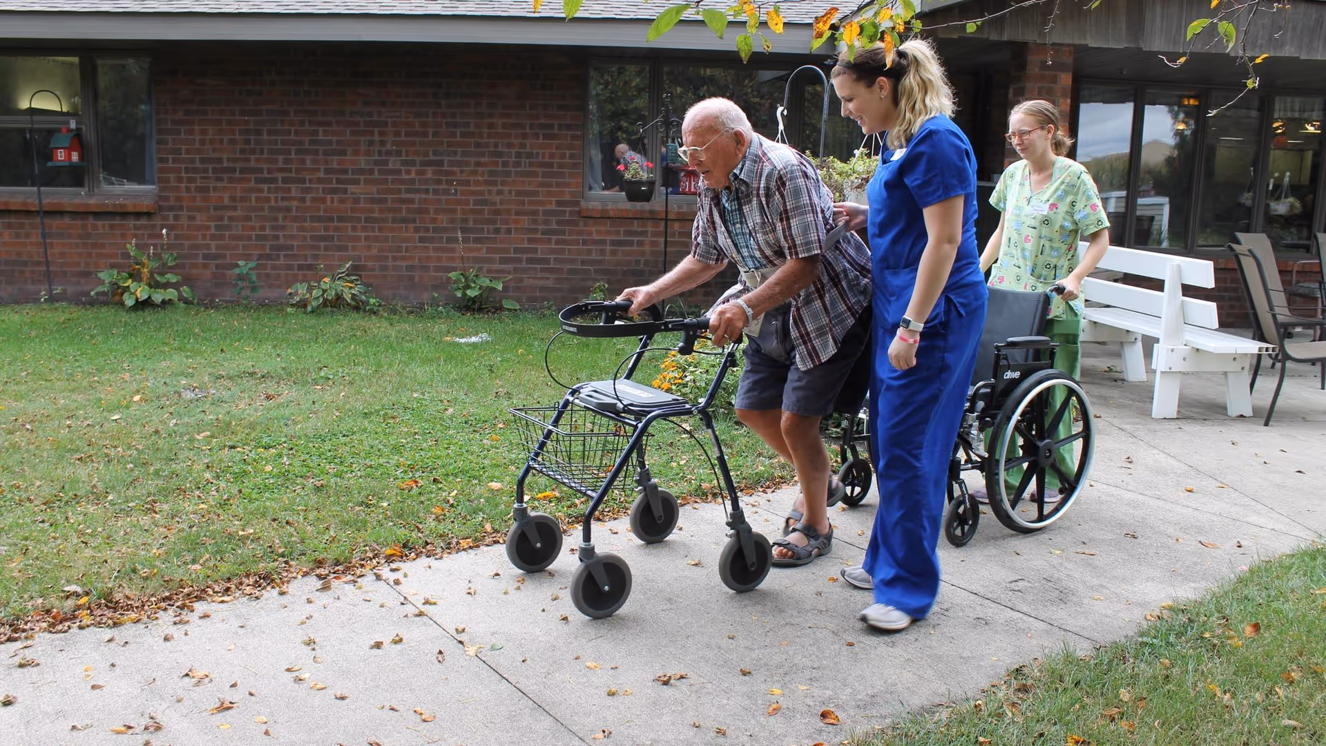 An elderly man using a walker is assisted by two female caregivers outside on a paved pathway next to a grassy area and a brick building. One caregiver is wearing blue scrubs and walking beside him, while the other caregiver in green scrubs is pushing an empty wheelchair behind them.
