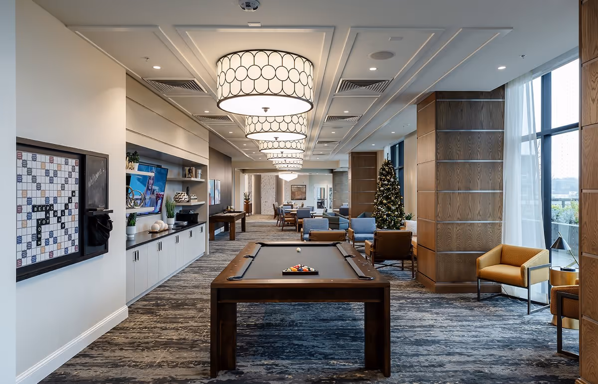 A spacious and modern common area in a senior living facility featuring a pool table in the center, comfortable seating with armchairs and sofas, a decorated Christmas tree near large windows with sheer curtains, and a wall-mounted TV. The room is well-lit with large circular ceiling lights and has a carpeted floor with a patterned design. A Scrabble board is mounted on the left wall.