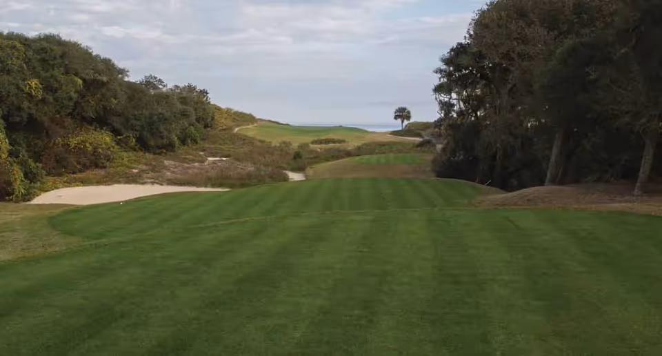 A well-maintained golf course fairway with green grass, sand bunkers on the left, surrounded by trees and bushes, under a cloudy sky.