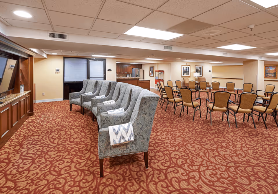 A spacious common area in a senior living facility featuring a row of patterned armchairs with folded blankets on them facing a large flat-screen TV mounted on a wooden entertainment unit. Behind the chairs, there are multiple rows of beige chairs arranged in a semi-circle, with framed artwork on the walls and a popcorn machine near a kitchenette area in the background. The room has a red patterned carpet and a drop ceiling with fluorescent lighting.