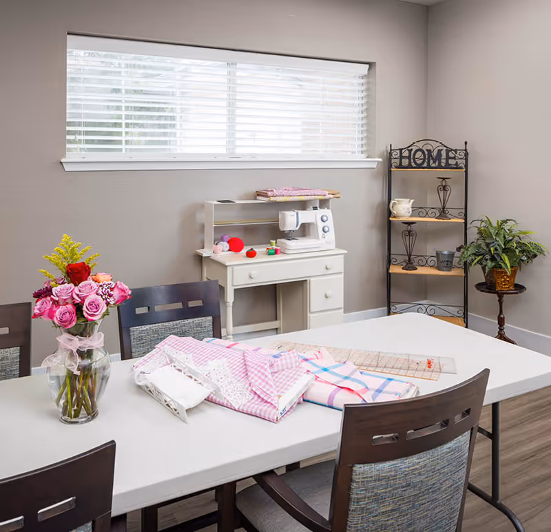 Bright interior activity room with a long table and chairs covered in fabric and sewing supplies, a sewing machine on a small desk, shelving, and a vase of flowers under a window.