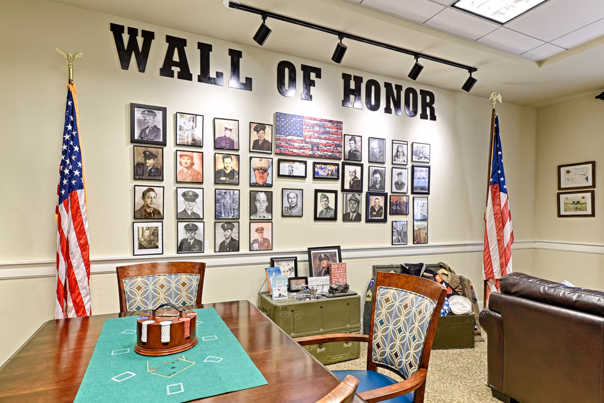 A room with a 'Wall of Honor' displaying framed photographs of military personnel. Two American flags stand on either side of the wall. In front of the wall is a table with a green felt poker mat and a poker chip holder. There are patterned chairs around the table and a brown leather couch to the right. Various military memorabilia are displayed on green storage trunks beneath the photos.