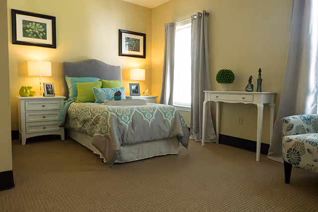 A cozy bedroom with a single bed featuring a patterned comforter and multiple pillows in shades of green and blue. There are two white nightstands on either side of the bed, each with a lamp and framed photos. The room has beige walls, carpeted floor, two windows with gray curtains, a white desk with decorative items, and a patterned armchair.