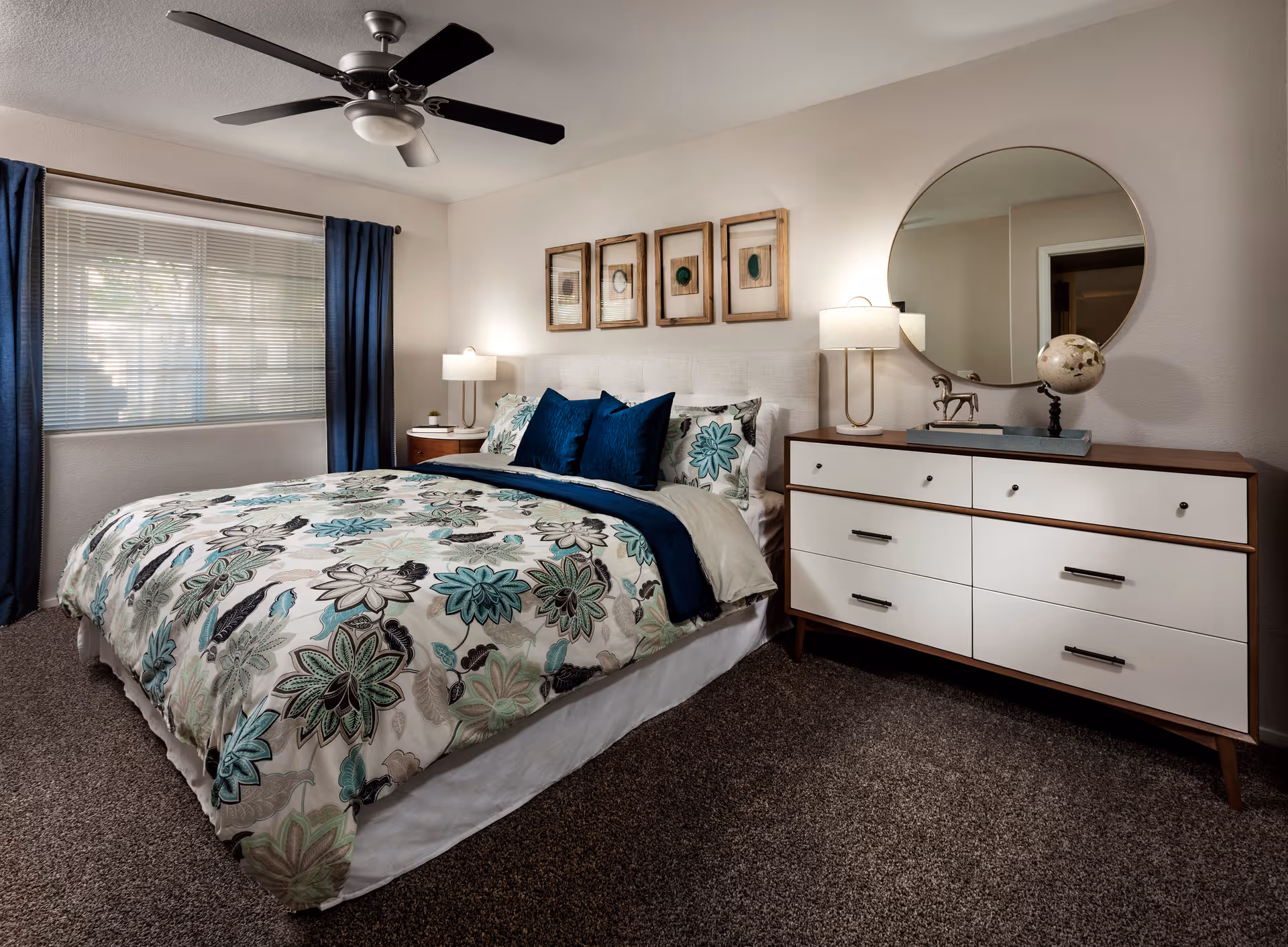 A neatly made bed with a floral patterned comforter and blue accent pillows in a bedroom. The room features a ceiling fan, a window with blue curtains, a wooden dresser with white drawers, a round mirror above the dresser, and two table lamps on either side of the bed. Four framed artworks hang above the bed's headboard.