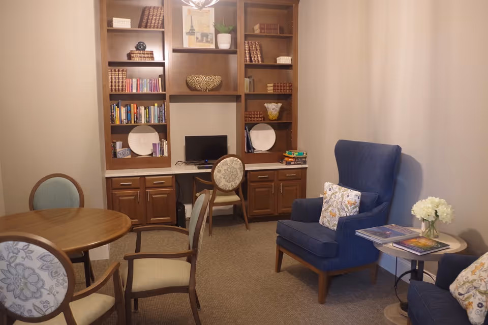 A cozy room with a wooden round table surrounded by four upholstered chairs, a built-in wooden bookshelf filled with books and decorative items, a small computer desk with a chair, and a blue armchair with a floral pillow next to a small round side table holding flowers and books.