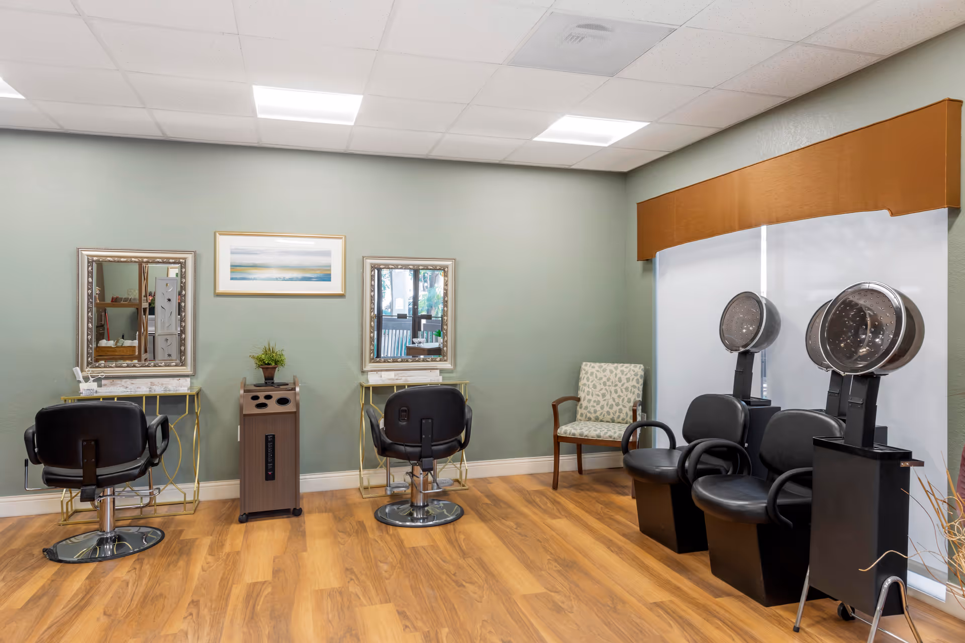 Interior view of a salon area with two black salon chairs in front of mirrors on a light green wall. There are two hair drying stations with black chairs and hooded hair dryers on the right side, and a patterned armchair near a window with a brown valance. The floor is wood, and the ceiling has recessed lighting.