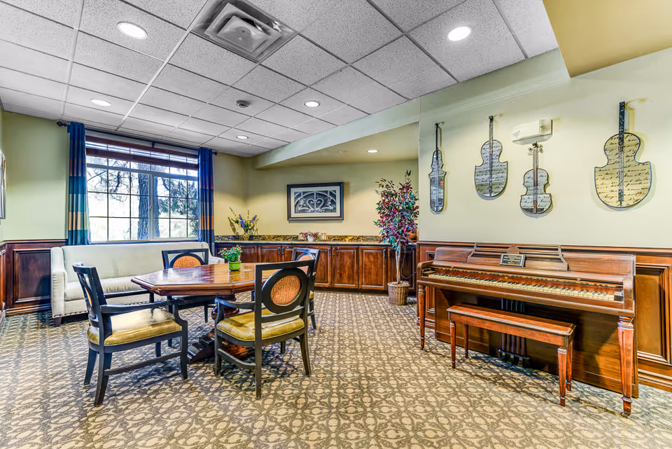 A cozy senior living common area with a wooden table surrounded by four chairs and a cushioned bench near a window with blue and orange curtains. On the right side, there is a wooden piano with a matching bench and decorative violin-shaped wall art above it. The room has patterned carpet flooring, wood paneling on the lower walls, and a ceiling with recessed lighting.