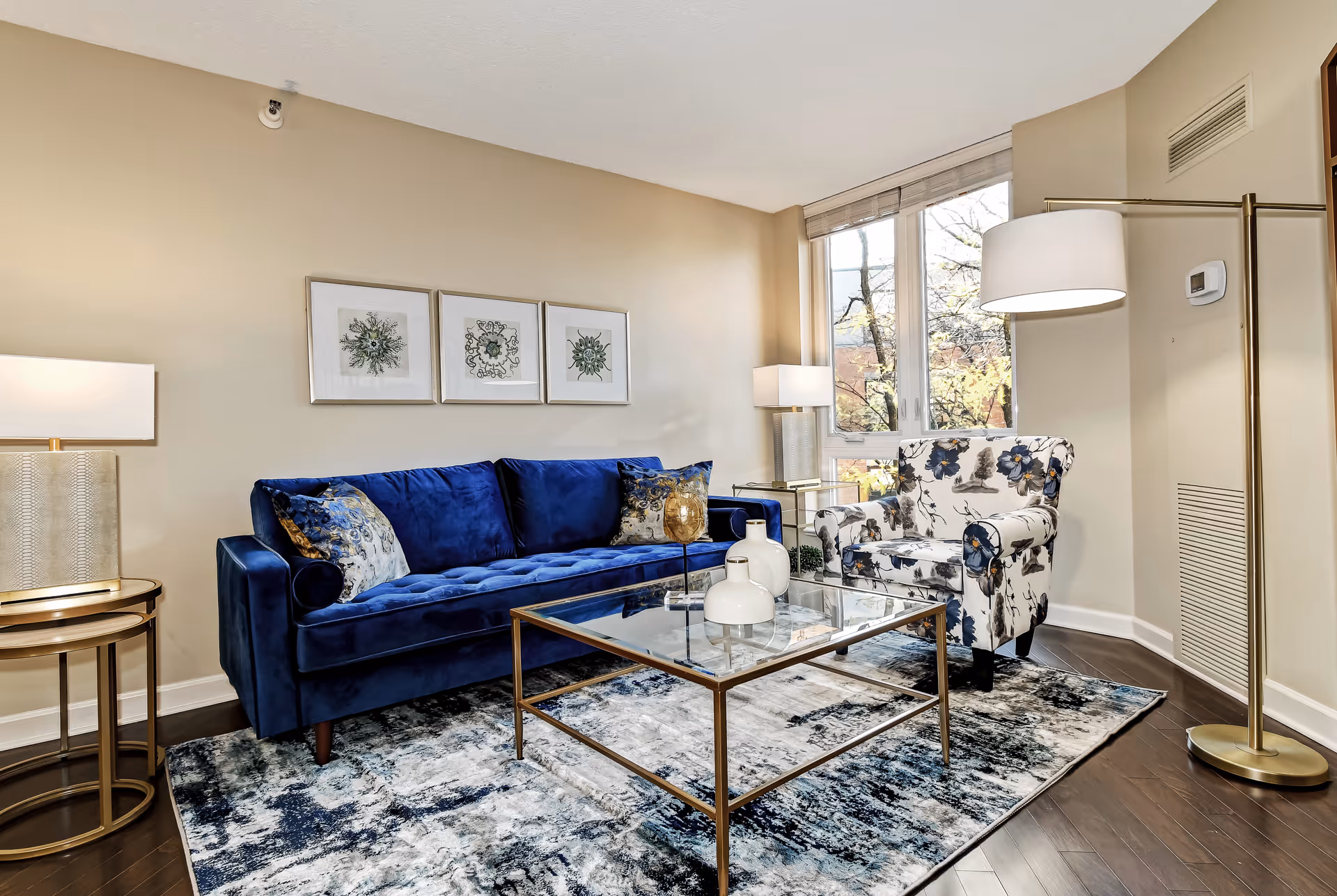 A cozy living room with a deep blue velvet sofa adorned with patterned cushions, a glass coffee table with decorative vases, a floral upholstered armchair, a floor lamp with a white shade, and a side table with a lamp. The room has light beige walls, three framed botanical prints above the sofa, and a large window letting in natural light.