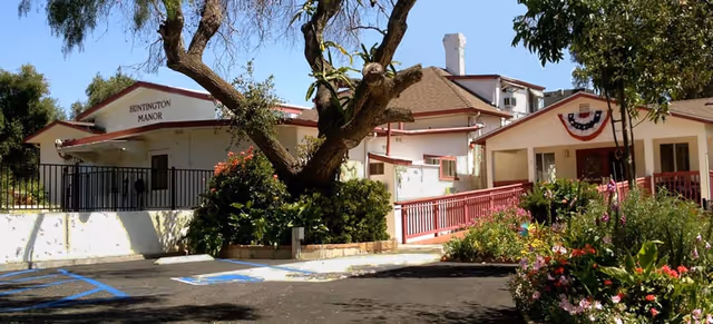 Exterior view of Huntington Manor, a single-story building with a sloped roof, surrounded by trees and flowering plants. There is a parking area with handicap spaces in front, and a ramp leading to the entrance. The building is decorated with a patriotic banner.