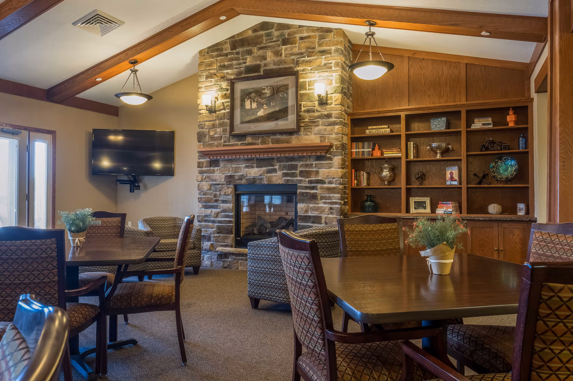 A cozy senior living common area with wooden beams on the ceiling, a stone fireplace with a framed picture above it, a wall-mounted flat screen TV, and built-in wooden shelves filled with books and decorative items. Several wooden tables and cushioned chairs are arranged around the room, with small potted plants on the tables.