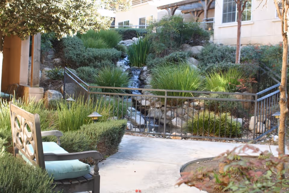 Outdoor garden area with a wooden bench featuring green cushions, surrounded by lush greenery and plants. A small waterfall flows over rocks in the background, with a building partially visible behind the garden. There is a metal railing along the pathway near the waterfall.