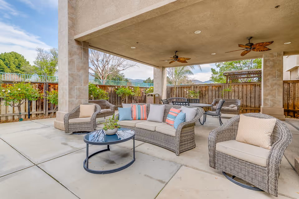 Covered outdoor patio with wicker sofas and chairs, a round coffee table, ceiling fans, and a fenced yard.