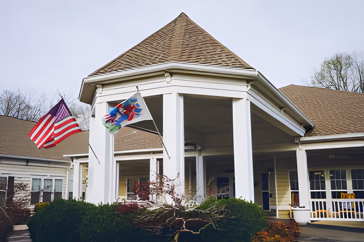 Exterior view of a senior living facility building with a peaked roof and white pillars. Two flags, including an American flag, are mounted on the front porch area. Green bushes and some trees are visible around the building.