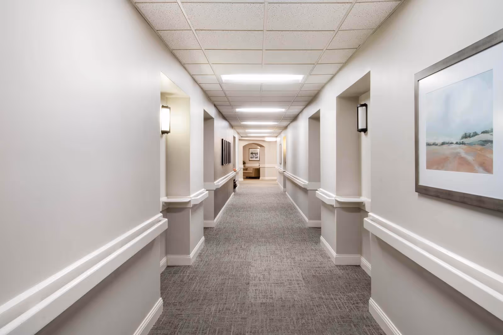 A long, well-lit hallway in a senior living facility with light gray walls, carpeted floor, handrails on both sides, and framed artwork on the walls. At the end of the hallway, there is a seating area with a couch and a picture on the wall.