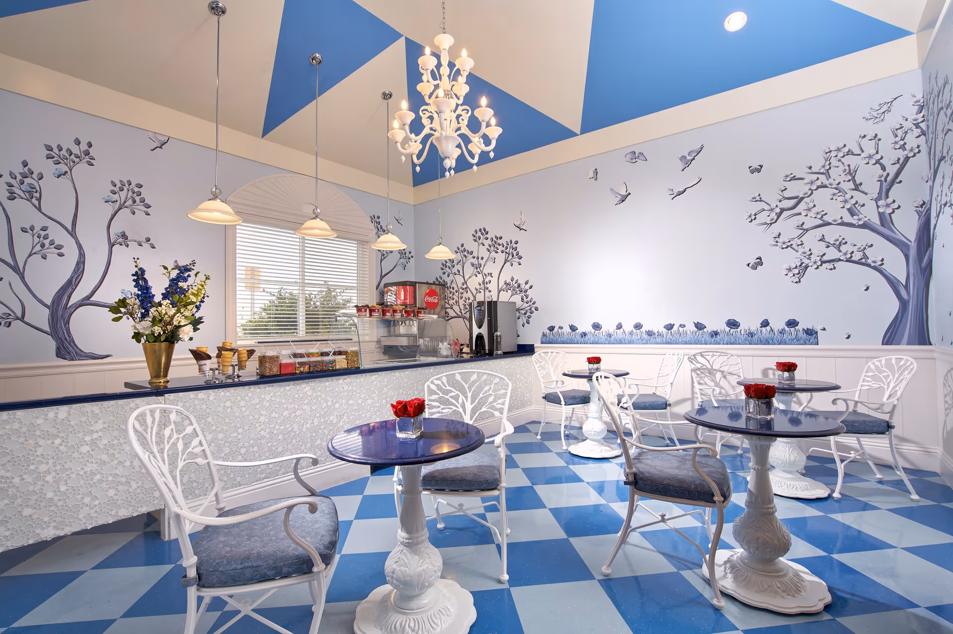 A bright and charming dining area with blue and white checkered floor tiles, white metal chairs with blue cushions, and round blue tables each adorned with a small vase of red flowers. The walls are decorated with artistic murals of trees and birds, and the ceiling features a blue and beige geometric pattern with a white chandelier and hanging pendant lights. A counter with a coffee machine, snacks, and a Coca-Cola dispenser is visible under a window with blinds.