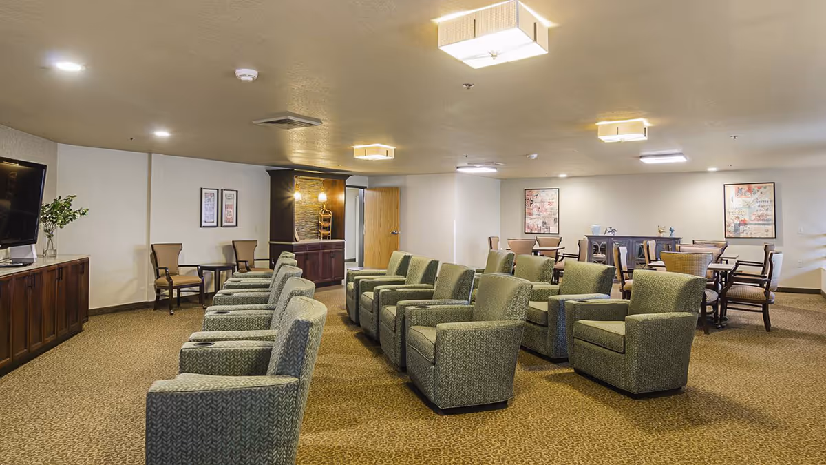 A spacious common area with two rows of green upholstered armchairs facing a large flat-screen TV mounted on a wooden cabinet. In the background, there are several tables with chairs, framed artwork on the walls, and a small counter with pendant lights. The room has beige walls, carpeted flooring, and ceiling lights.