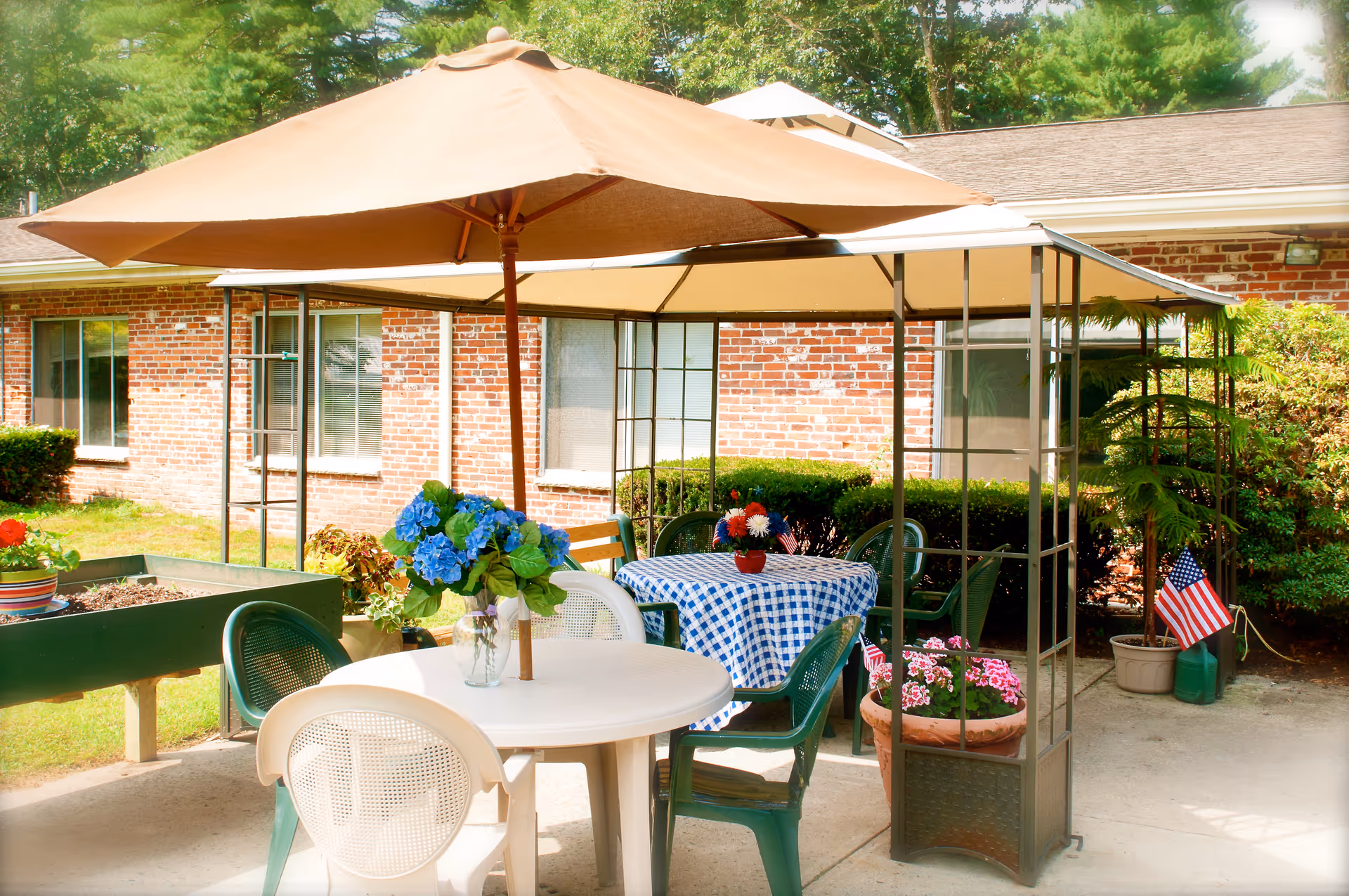 Outdoor patio area at Sudbury Pines Extended Care with tables and chairs under large umbrellas. One table has a blue and white checkered tablecloth with a small flower arrangement, and another table has a vase with blue flowers. There are potted plants and small American flags around the patio, with a brick building and greenery in the background.