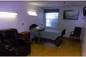 A small room with a single bed, a bedside table, a reclining chair, a rolling table, and two additional chairs near the window. The room has wooden flooring, a window with blinds, and a framed picture on the wall.