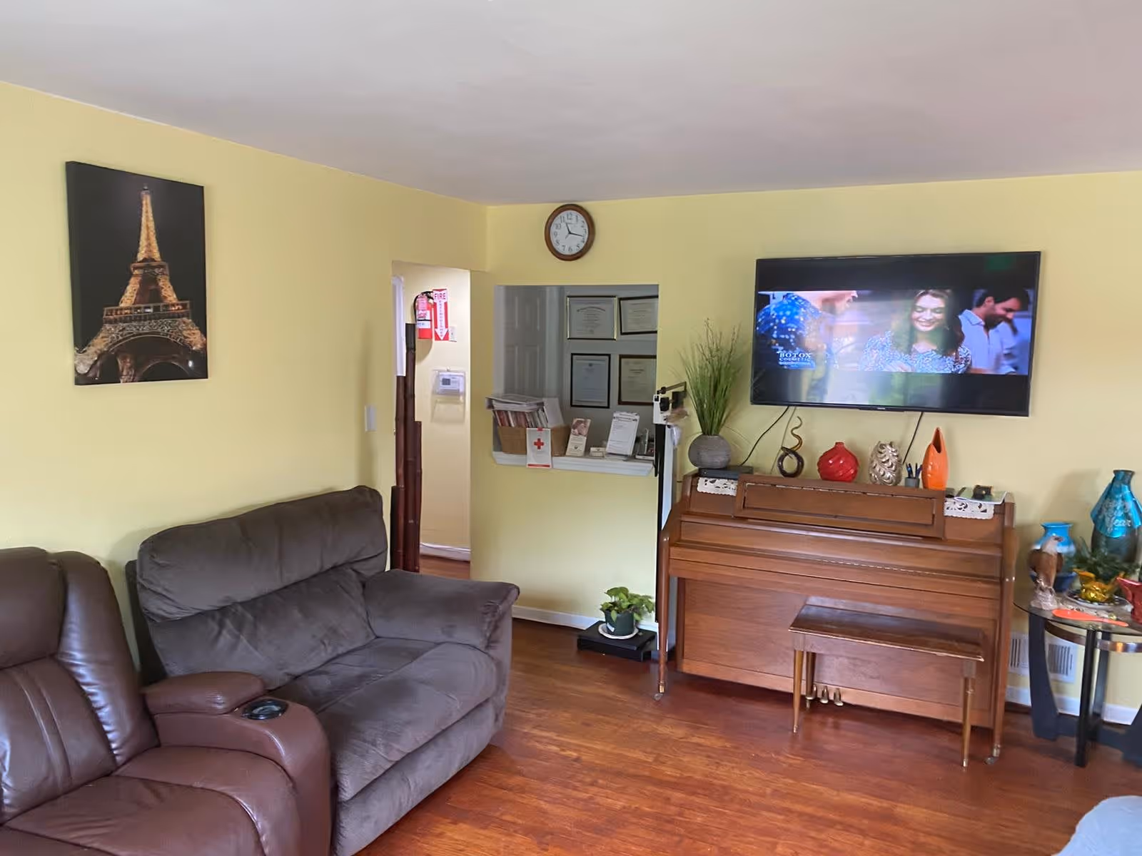 A cozy living room with yellow walls featuring a brown leather recliner and a matching brown sofa. On the wall, there is a framed picture of the Eiffel Tower and a clock above a small reception window. A wooden piano with a bench is placed against the wall under a mounted flat-screen TV showing a scene with people. Decorative items and plants are arranged on and around the piano.