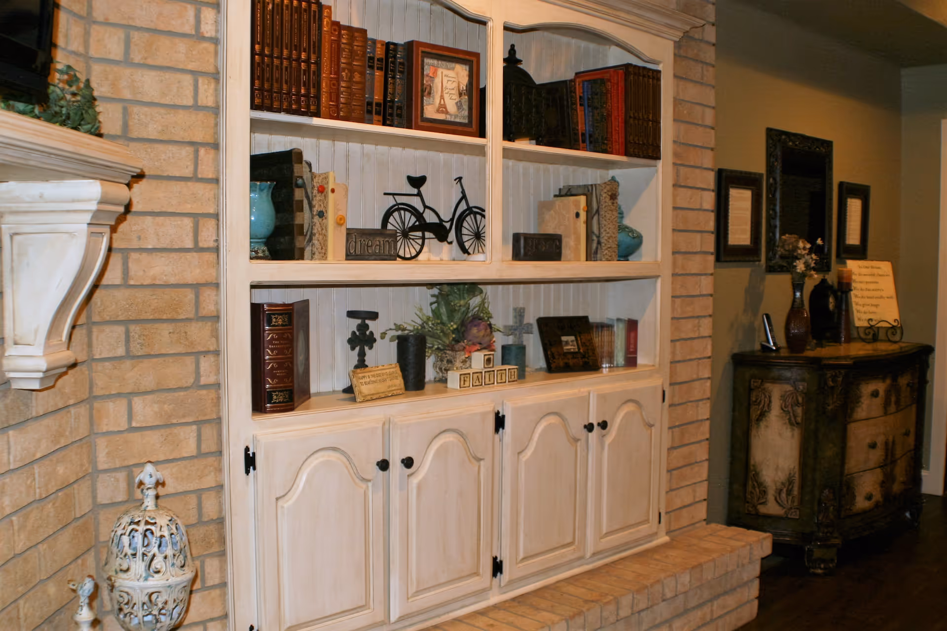 Interior view of a cozy living room area featuring a built-in white wooden bookshelf filled with books, decorative items including a small bicycle figurine, framed pictures, and blocks spelling 'FAITH'. The bookshelf is set against a brick wall with a white mantle on the left. To the right, there is a vintage-style wooden cabinet with decorative vases, framed pictures, and a sign with text, along with a mirror and framed artwork on the wall.