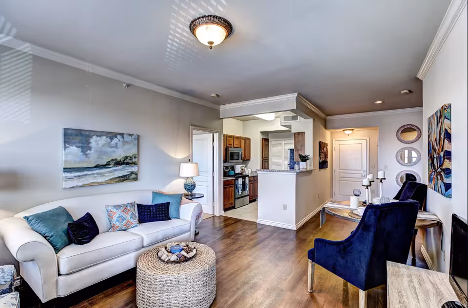 A cozy living room with a white sofa adorned with colorful pillows, a round woven ottoman, two dark blue armchairs, a wooden side table with candles, and a wall decorated with paintings and round mirrors. The room has wooden flooring and leads into a kitchen area with wooden cabinets and stainless steel appliances.