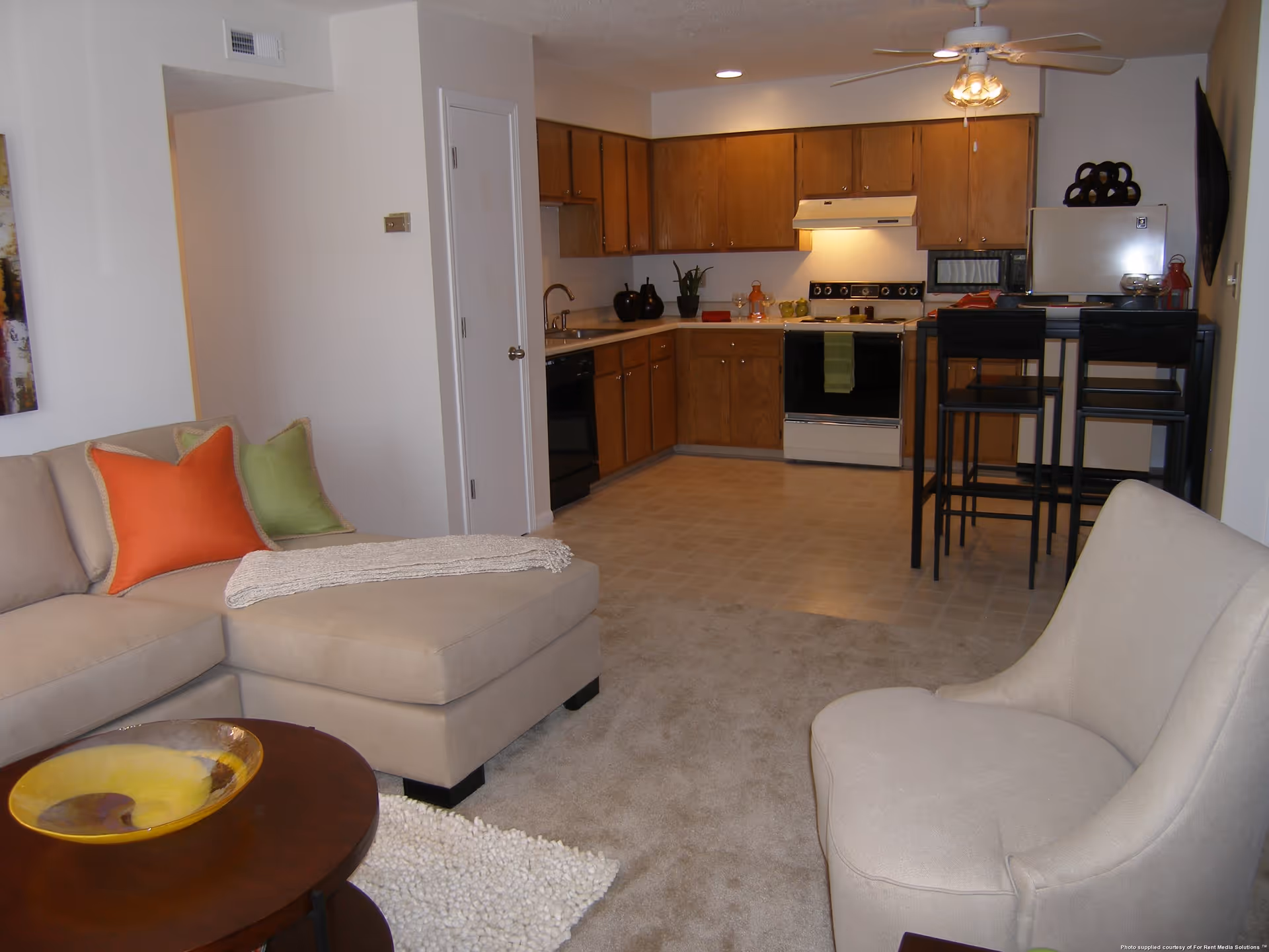 Interior view of a senior living facility apartment showing a living room area with a beige sectional sofa adorned with orange and green pillows, a beige armchair, and a round wooden coffee table with a decorative yellow bowl. The living room opens into a kitchen with wooden cabinets, a stove, a refrigerator, a microwave, and a small dining table with four black chairs. The space is carpeted in the living area and has vinyl flooring in the kitchen.