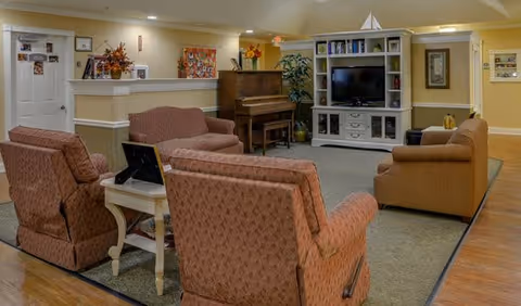 A cozy senior living common area with several upholstered armchairs and a loveseat arranged around a white side table. In the background, there is a wooden piano, a white entertainment center with a flat-screen TV, and decorative plants and artwork on the walls. The room has warm lighting and a welcoming atmosphere.