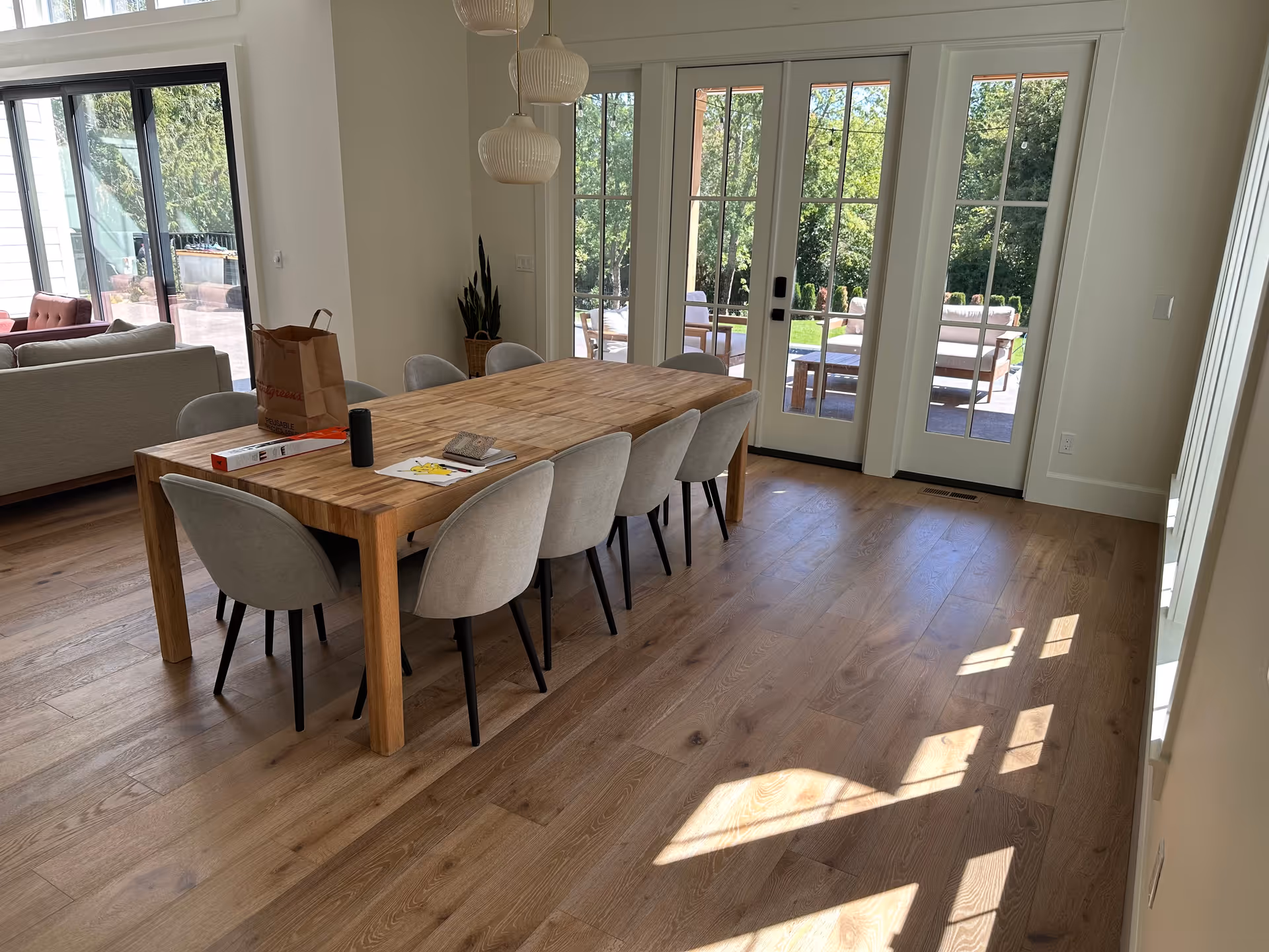 A bright dining room with a wooden table surrounded by eight gray upholstered chairs. On the table are a paper bag, a black cylindrical object, some papers, and a small book. The room has wooden flooring and large glass doors leading to an outdoor patio with seating and greenery visible outside. A gray sofa is partially visible to the left.