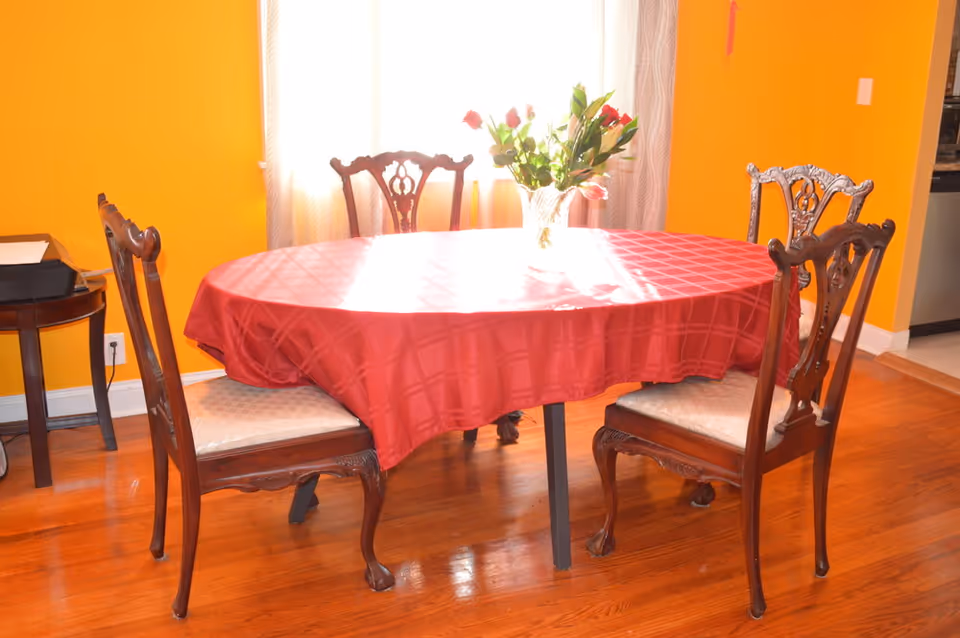 A dining room with a wooden oval table covered by a red tablecloth. Four wooden chairs with cushioned seats surround the table. A vase with flowers is placed in the center of the table. The walls are painted bright orange, and sunlight streams in through a window with light-colored curtains behind the table. A small side table with some items is visible to the left.