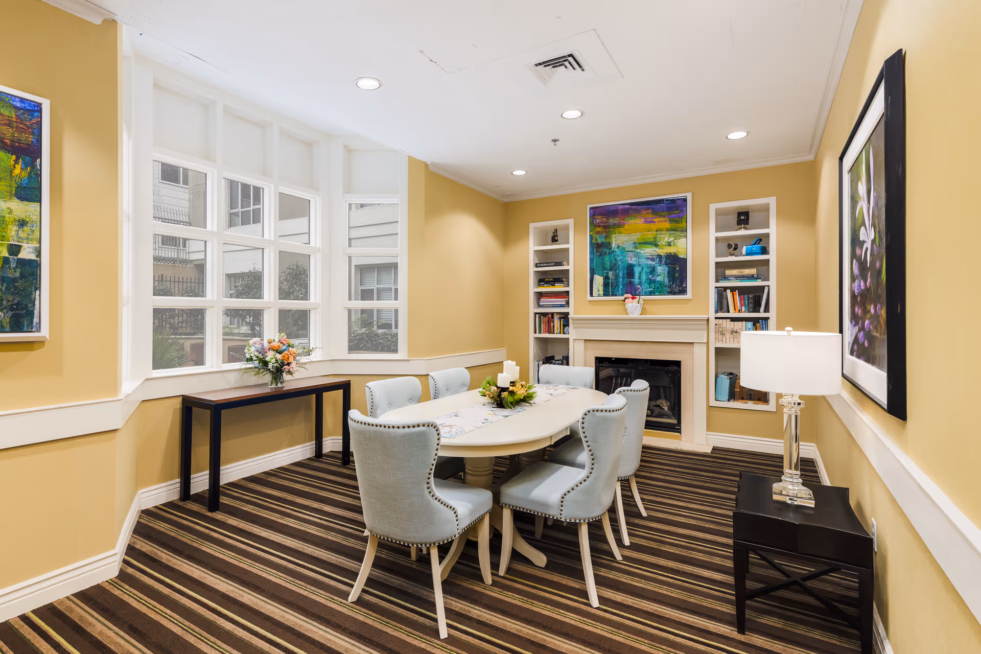 A cozy dining room with a white oval table surrounded by six light blue upholstered chairs. The room has yellow walls, a striped carpet, and large windows letting in natural light. There is a fireplace with built-in shelves on either side, decorated with books and colorful artwork above it. A small table with a flower arrangement is placed under the windows, and a side table with a lamp is on the right wall.