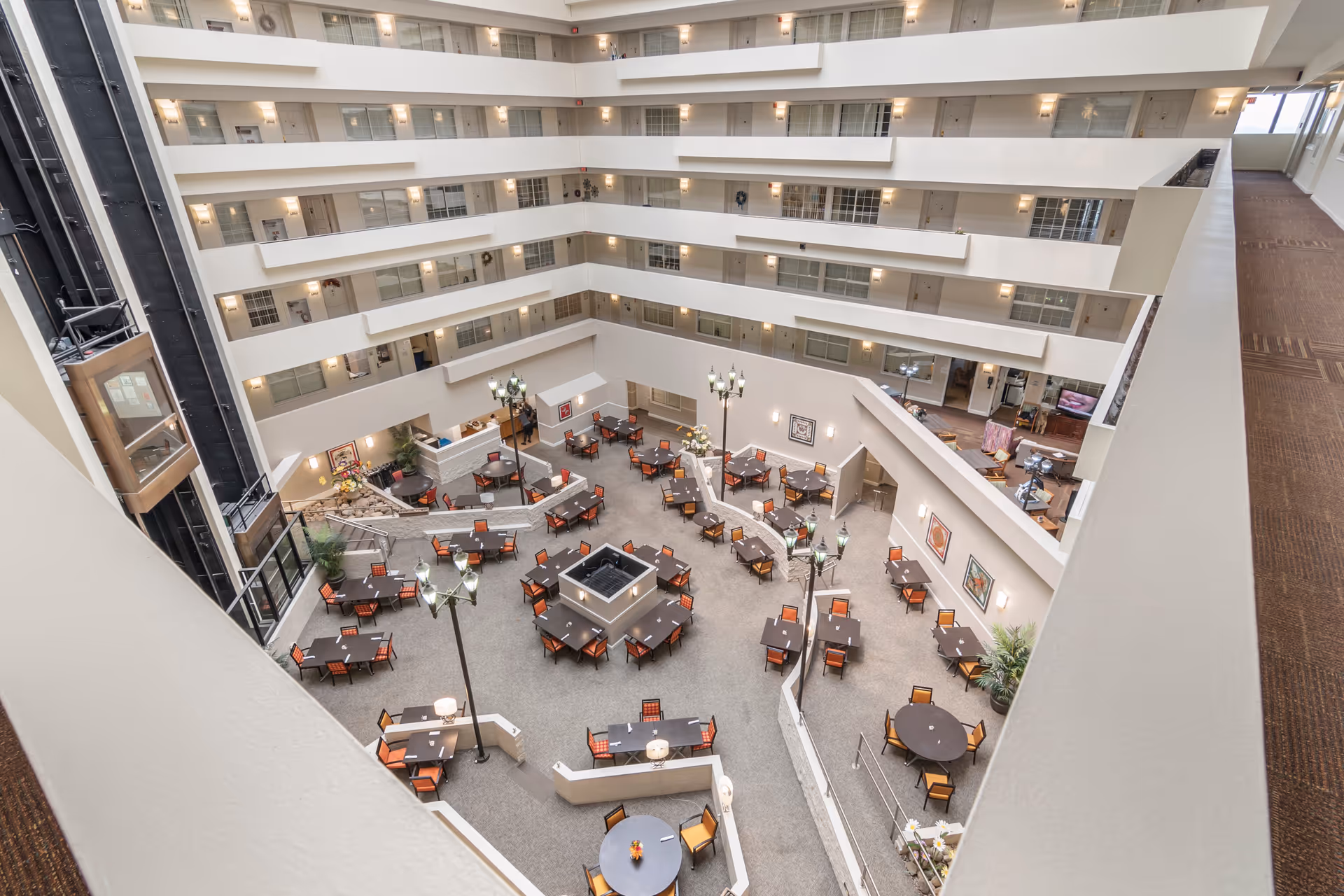 View from an upper floor overlooking a large indoor atrium area with multiple tables and chairs arranged for dining or socializing. The atrium is surrounded by several floors of rooms with balconies and glass elevators on the left side. The space is well-lit with tall lamp posts and decorated with framed artwork on the walls.