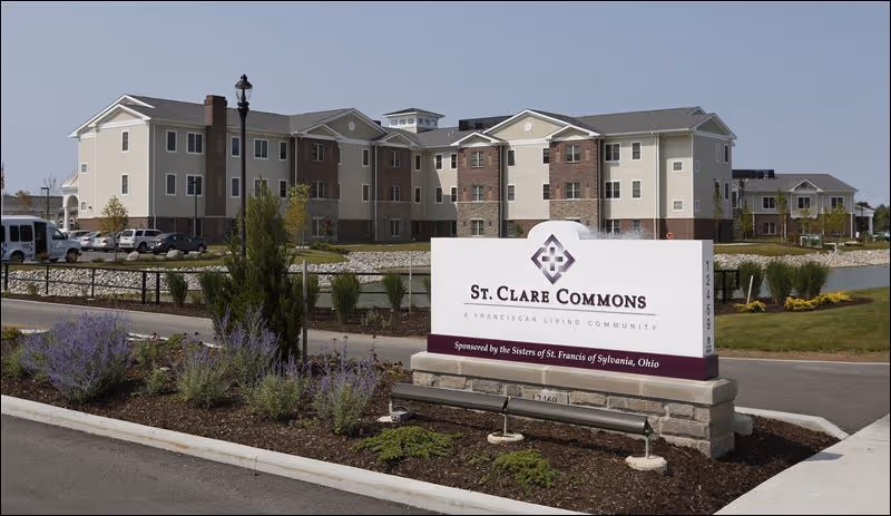 Exterior view of St. Clare Commons, a three-story senior living community building with beige and brick facade, surrounded by landscaped grounds and a parking area. A large sign in the foreground displays the facility name and sponsorship information.