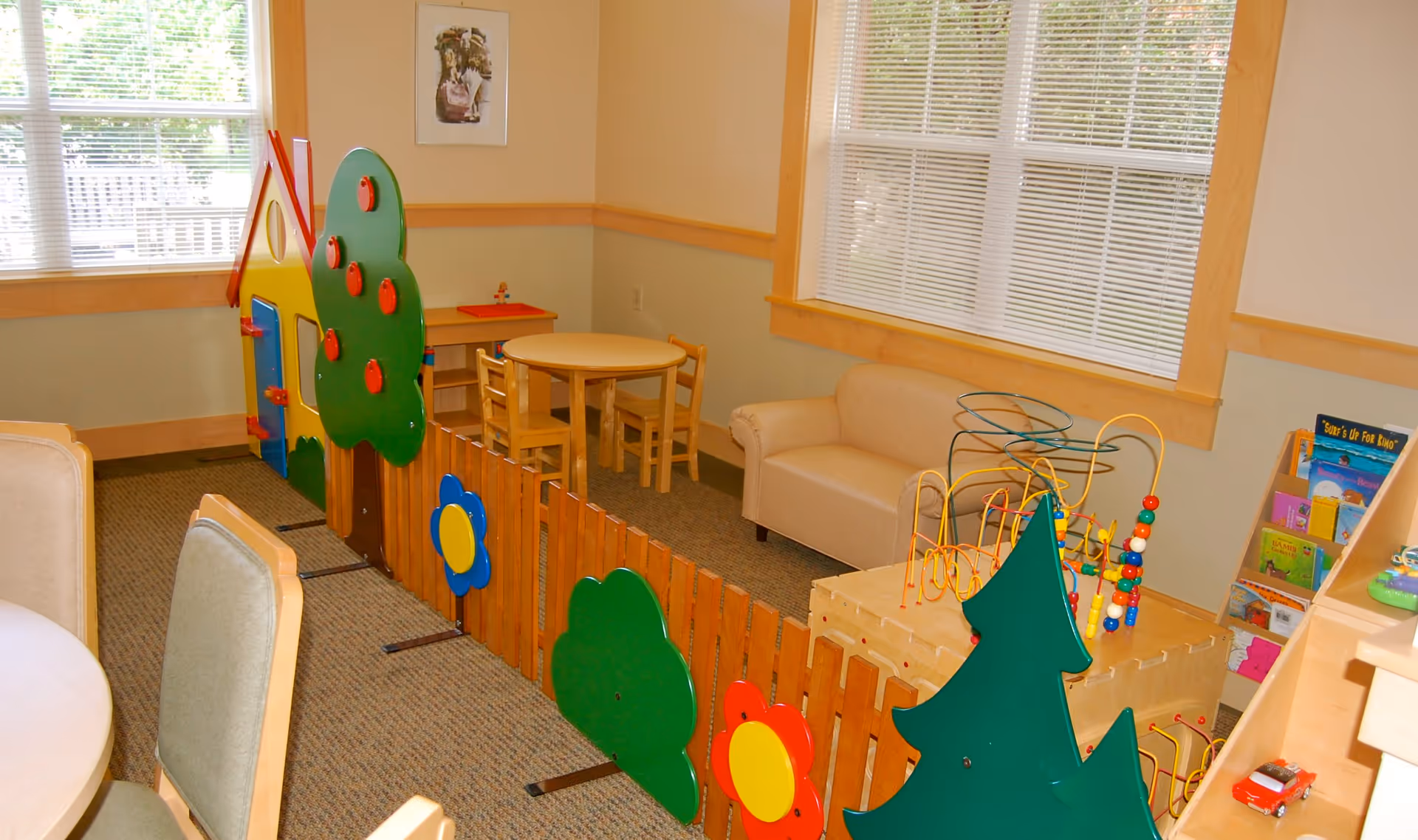 Interior common room with a fenced children's play area, small table and chairs, toys, and a couch by the windows.
