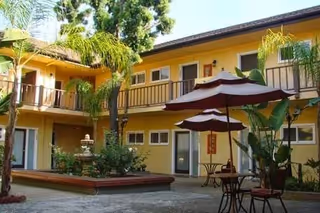 Outdoor courtyard area of a senior living facility with yellow two-story buildings surrounding it. The courtyard features a small fountain, palm trees, and patio tables with umbrellas.