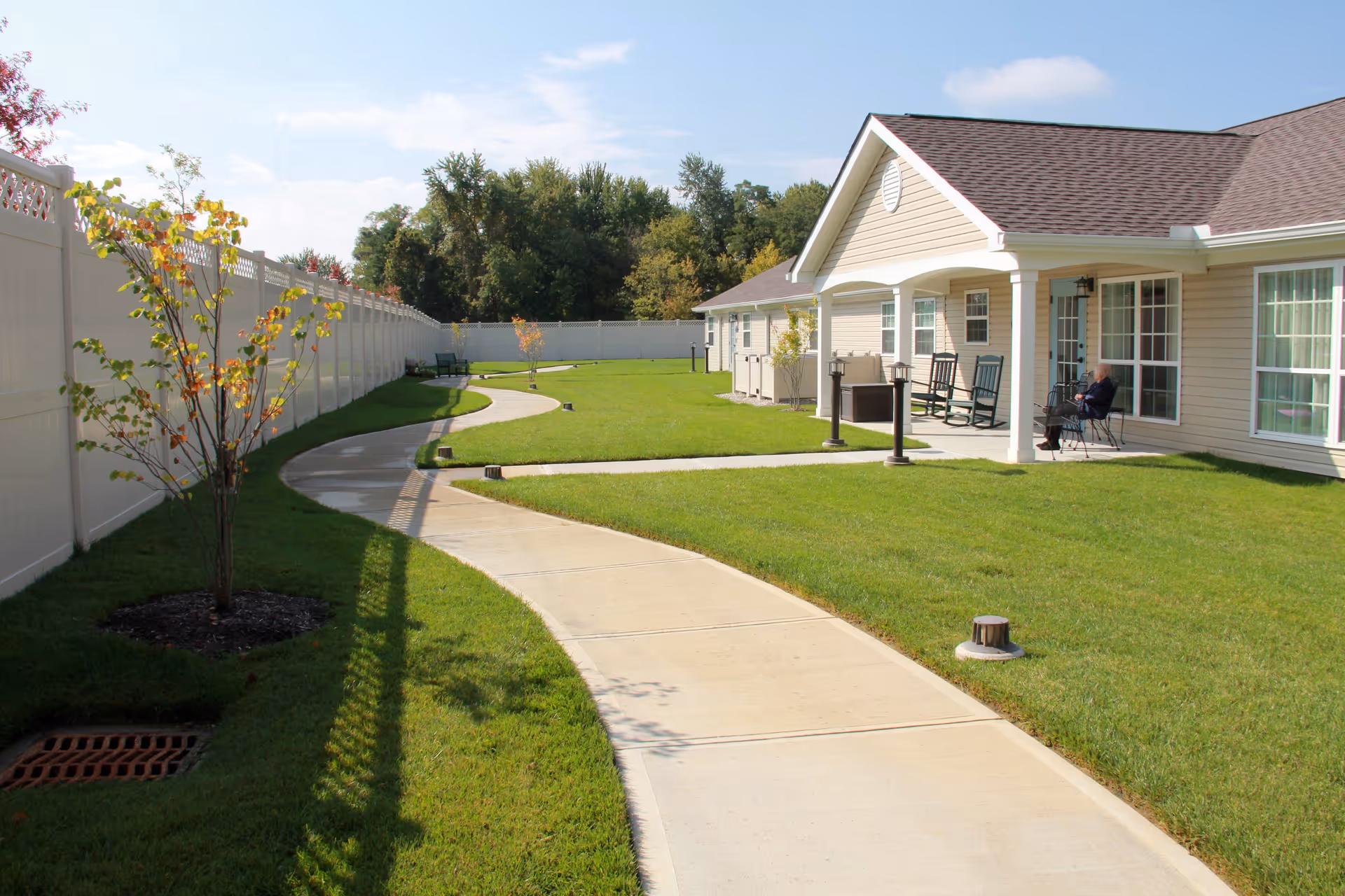 A sunny outdoor area at a senior living facility with a curved concrete walkway, green grass, small trees, and a beige building with a covered porch. On the porch, there are rocking chairs and a person sitting on one of the chairs. The area is enclosed by a white fence.