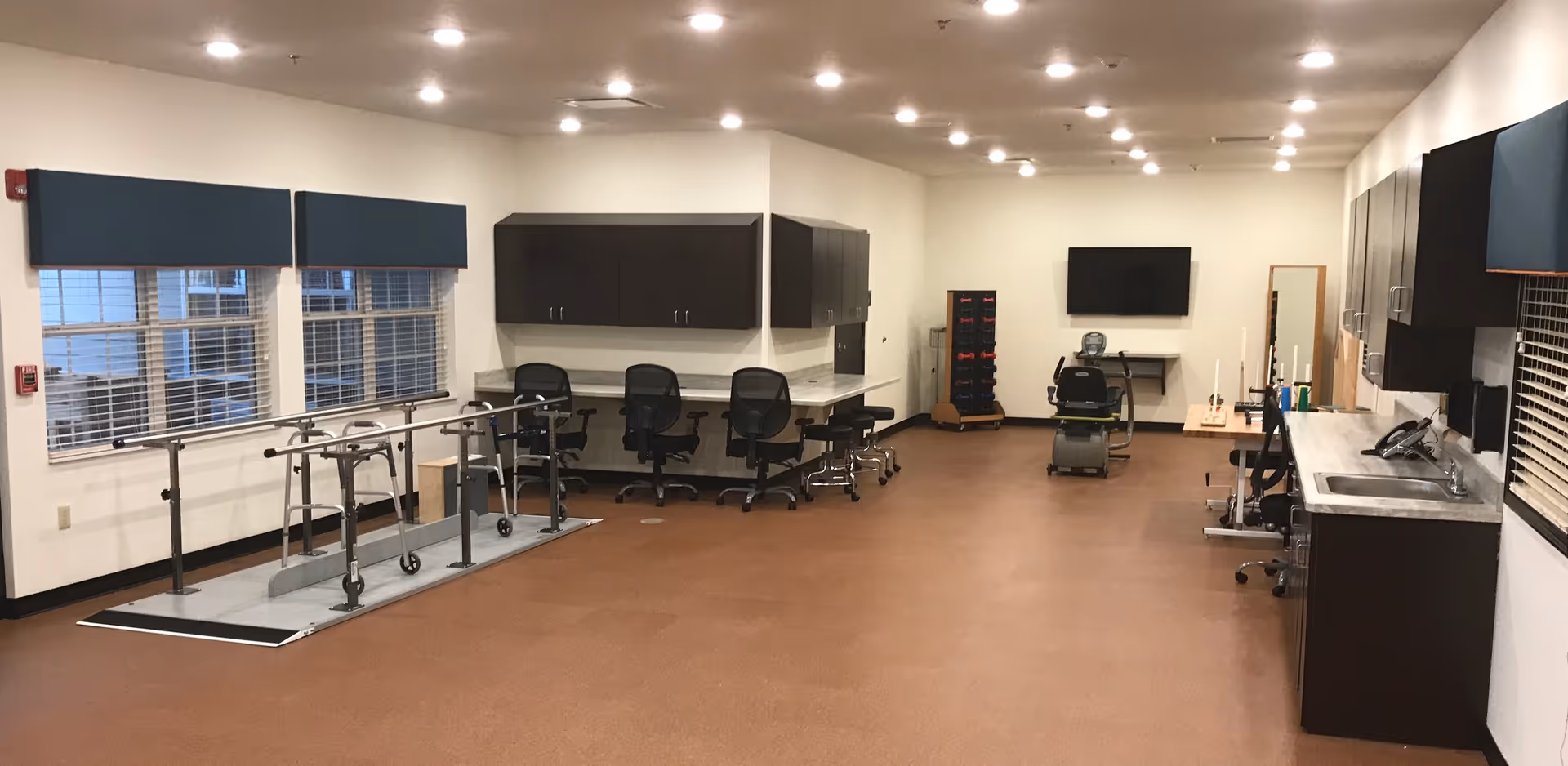 A spacious therapy or rehabilitation room with parallel bars for walking exercises on the left side, several office chairs and desks along the back wall, a TV mounted on the far wall, and a sink with cabinets on the right side. The room has large windows with blue valances and a brown floor.