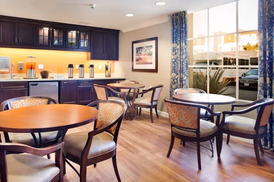 A bright dining area with round wooden tables and cushioned chairs with patterned upholstery. The room has a kitchenette with dark wood cabinets, a countertop with beverage dispensers, and a dishwasher. Large windows with blue patterned curtains let in natural light and show a view of a parked bus and car outside.
