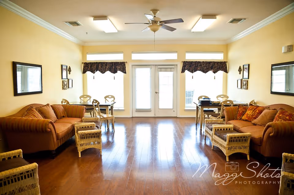 A bright and spacious living room with two orange sofas facing each other, each adorned with patterned cushions. Between the sofas are wicker coffee tables. The room has wooden flooring, cream-colored walls, and three large windows with dark valances letting in natural light. There are two small dining tables with chairs near the windows, and a ceiling fan with a light fixture is mounted on the ceiling. Wall decorations and mirrors are visible on the side walls.