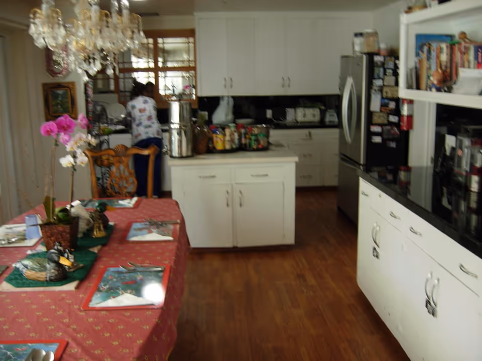 Interior view of a kitchen and dining area with wooden flooring. A dining table covered with a red tablecloth is set with placemats, utensils, and a potted orchid centerpiece. The kitchen has white cabinets, a central island with various items on it, and stainless steel refrigerator. A person is seen in the background near the sink area.