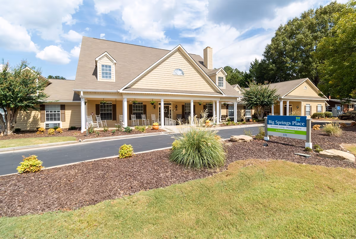 Exterior view of a single-story assisted living and memory care facility with beige siding, white columns, and a covered porch with rocking chairs. The building is surrounded by landscaped grounds with mulch, bushes, and trees under a partly cloudy sky. A sign in front reads 'Big Springs Place Assisted Living - Memory Care'.