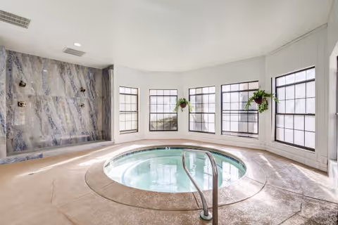 Indoor circular hot tub with clear water, surrounded by a textured concrete floor. The room has large windows letting in natural light, with two hanging potted plants near the windows. There is a marble or stone wall with multiple showerheads on the left side.