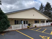 Exterior view of a single-story building with beige siding and a white railing along the front porch. There are several windows and doors visible, with some potted plants near the entrance. The parking lot in front has marked spaces, including a handicapped parking spot. Tall evergreen trees are visible in the background under a cloudy sky.