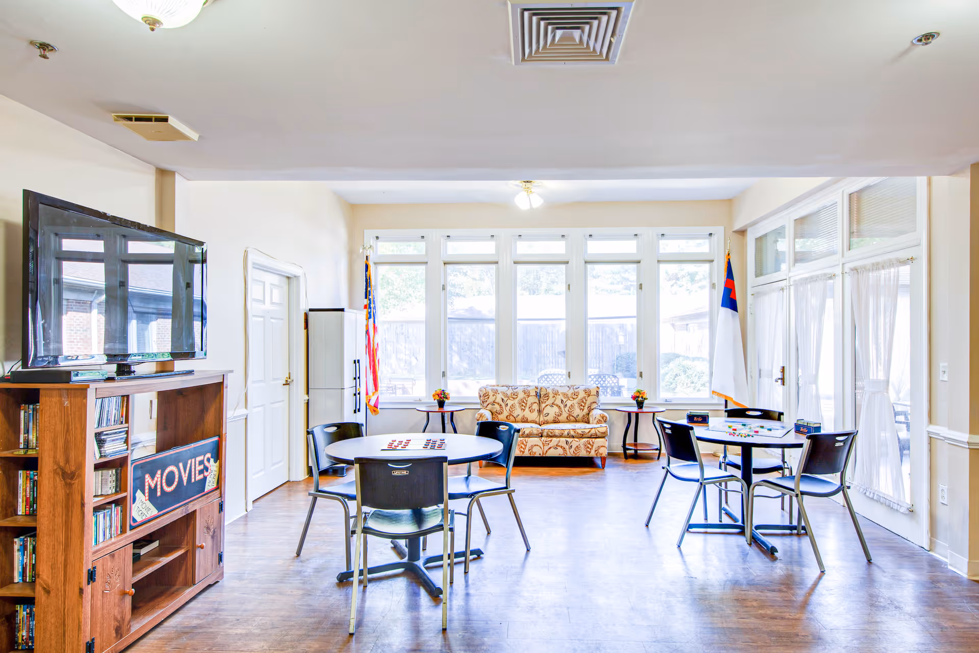 Bright community lounge with round game tables and chairs, a TV cabinet labeled 'MOVIES', and a patterned sofa in front of large windows.