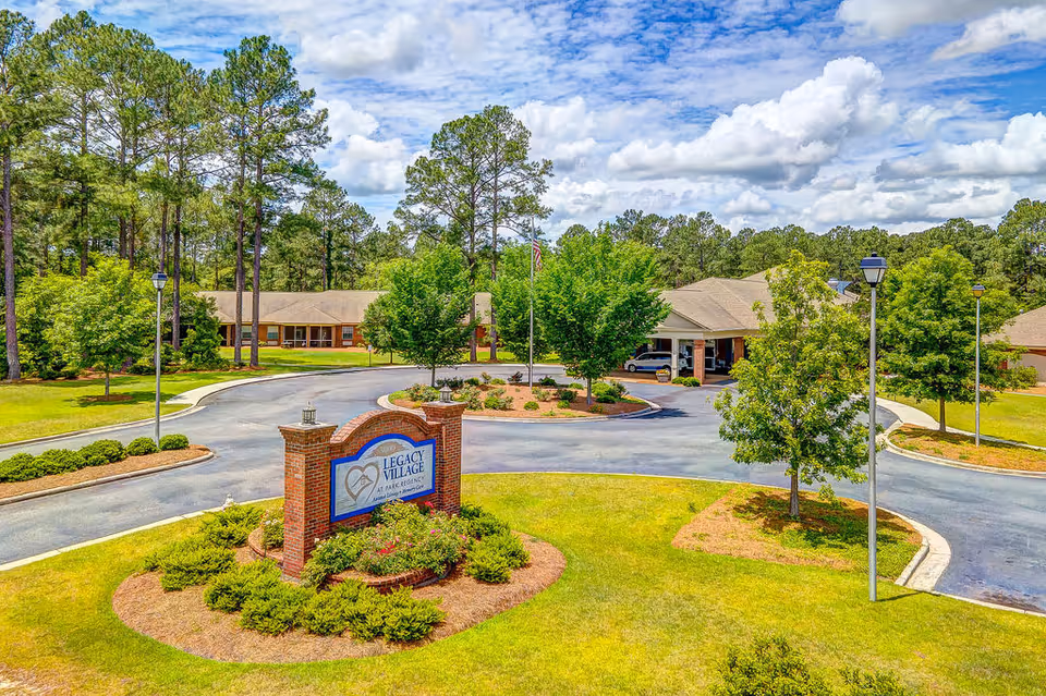 Entrance area of Legacy Village at Park Regency senior living facility with a brick sign surrounded by greenery, a circular driveway, trees, and buildings in the background under a partly cloudy sky.