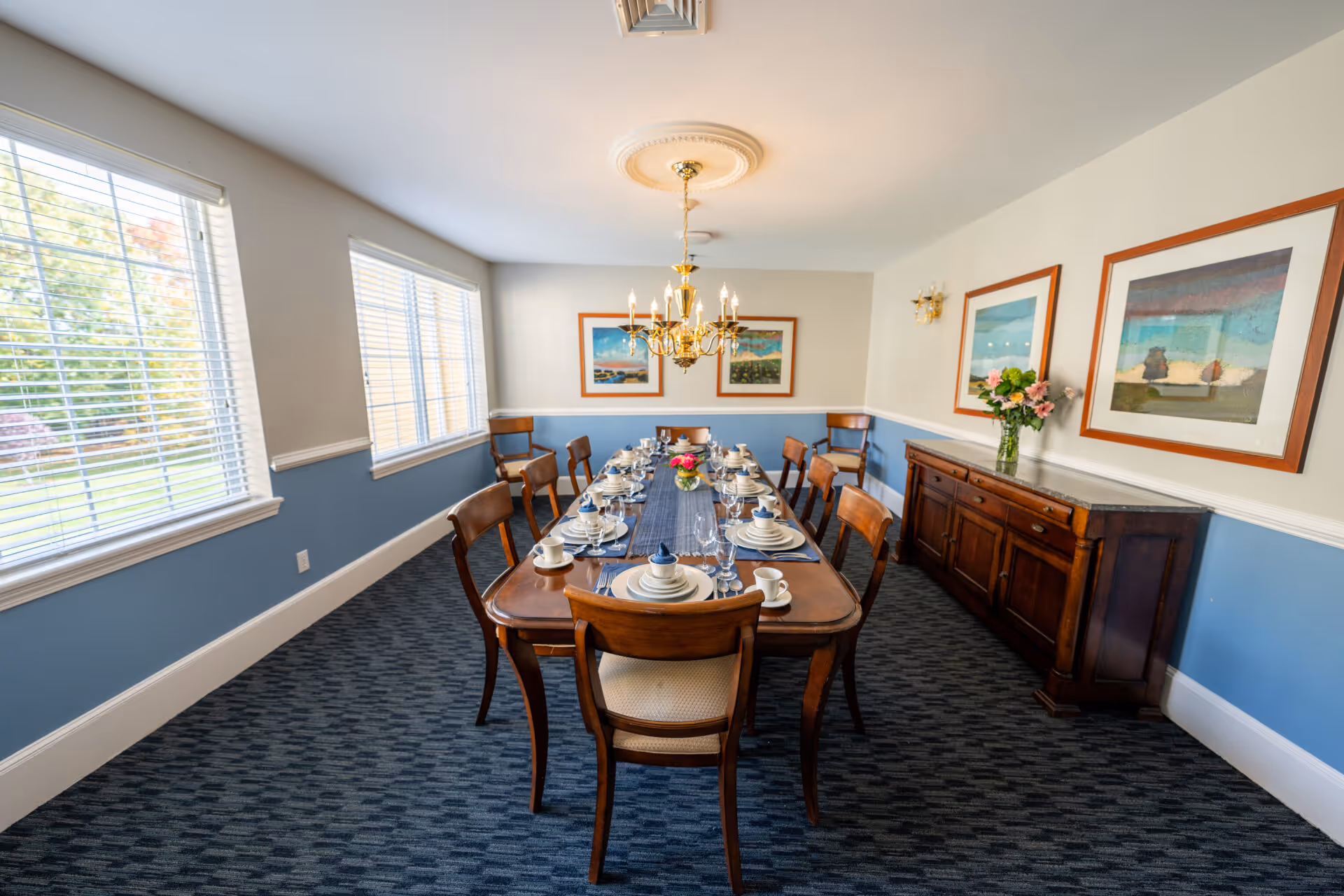 A formal dining room with a long wooden table set for a meal with plates, cups, glasses, and a centerpiece of flowers. The room has blue and white walls, carpeted floor, large windows with blinds letting in natural light, a chandelier hanging above the table, framed artwork on the walls, and a wooden sideboard with a vase of flowers.
