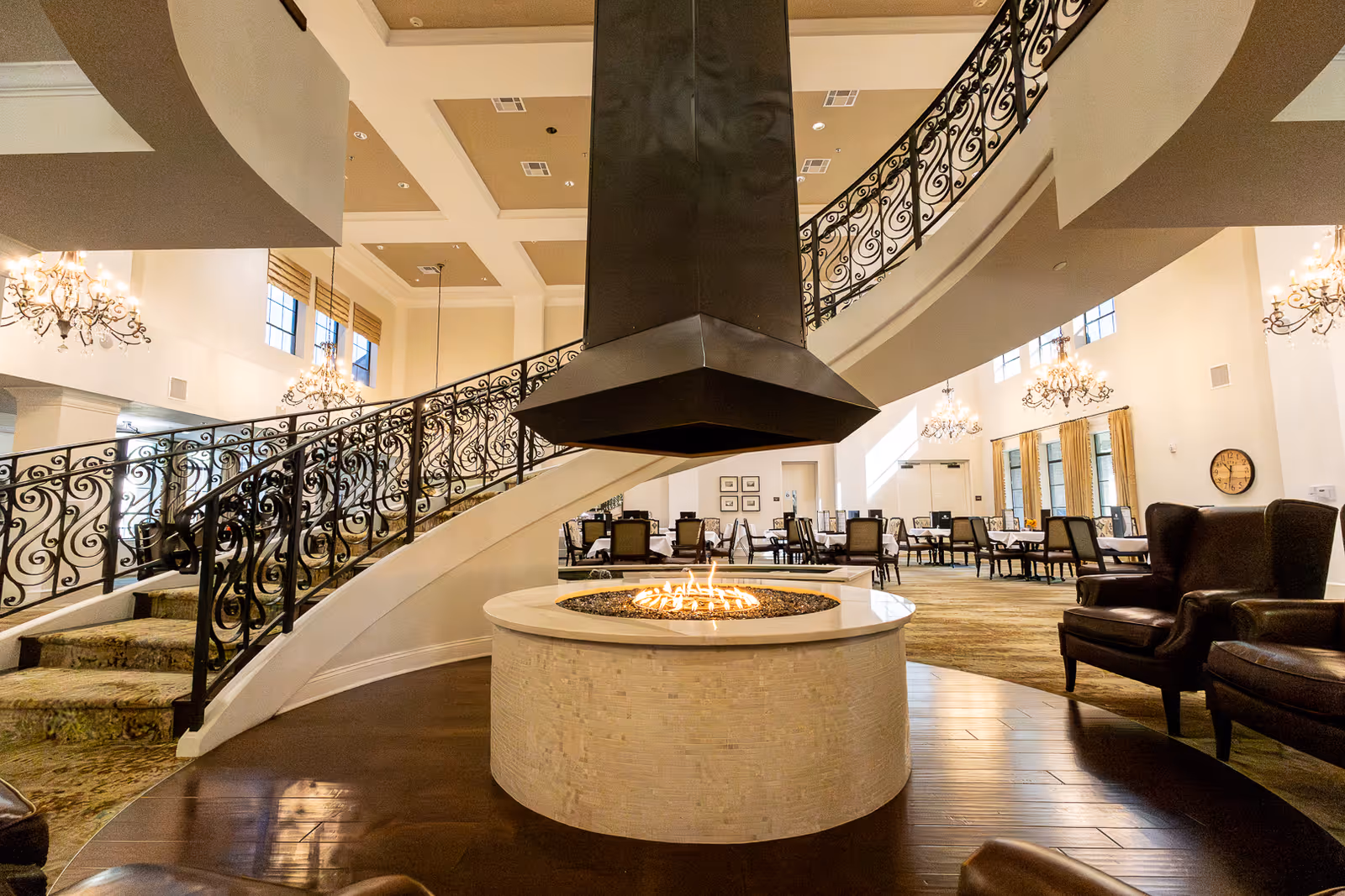 Spacious senior living facility common area with a central round fireplace, surrounded by comfortable chairs. A curved staircase with ornate black railings ascends to an upper level. The room is well-lit with large windows and elegant chandeliers, and there are multiple dining tables and chairs in the background.